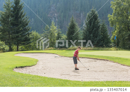 Boy standing in sand bunker holding golf club lining up golf ball at flagstick, copy space 135407014