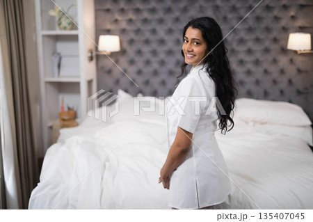 Indian woman standing beside bed with white linens, tufted headboard and lamps at hotel, copy space 135407045