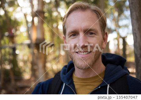 Bearded man standing in sunlit woods wearing navy hoodie and mustard shirt carrying black backpack Bearded man standing in sunlit woods wearing navy hoodie and mustard shirt carrying black backpack 135407047