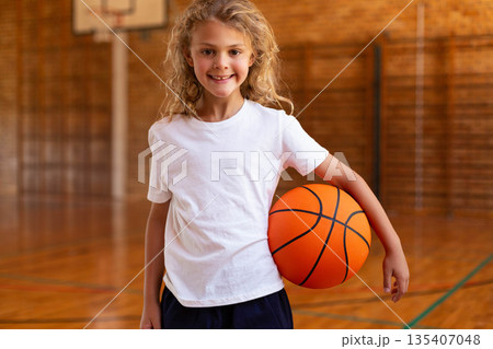 Child girl wearing sportswear holding basketball and smiling on gym court with hoop and lines Child girl wearing sportswear holding basketball and smiling on gym court with hoop and lines 135407048