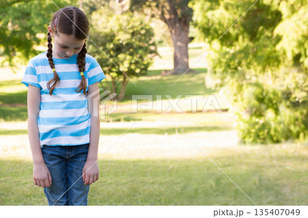 Girl standing on sunlit grassy lawn in park with braided pigtails looking down, copy space 135407049