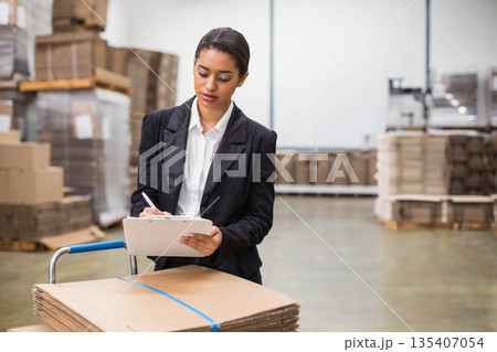 Woman leaning on trolley with cardboard cartons and writing on clipboard near pallets in warehouse Woman leaning on trolley with cardboard cartons and writing on clipboard near pallets in warehouse 135407054