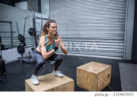 Mid adult woman performing box squat on plyometric box at gym with mirror showing squat rack 135407055