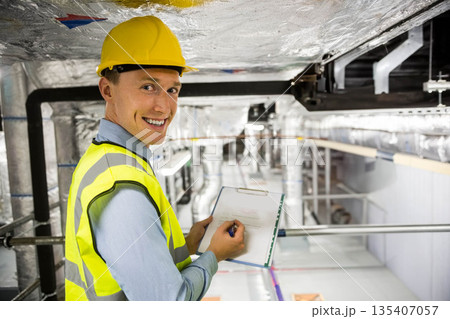 Male engineer wearing helmet, vest checking ducts in hall holding clipboard, pen, copy space Male engineer wearing helmet, vest checking ducts in hall holding clipboard, pen, copy space 135407057