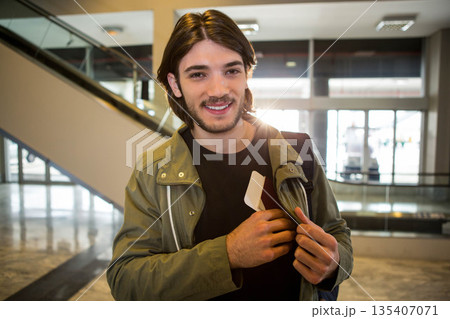 Man removing passport from green jacket pocket while standing in airport terminal carrying backpack 135407071