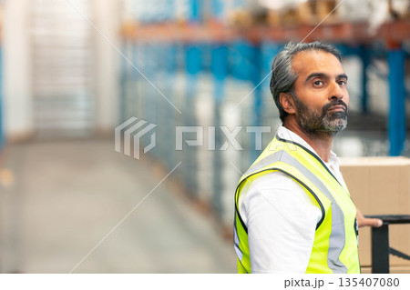 Middle-aged warehouse worker pushing dolly loaded with box wearing safety vest in aisle, copy space 135407080