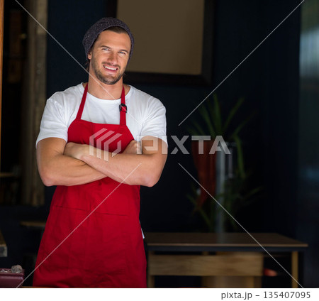Male barista wearing red apron and knit beanie, crossing arms, smiling behind wooden table at café Male barista wearing red apron and knit beanie, crossing arms, smiling behind wooden table at café 135407095