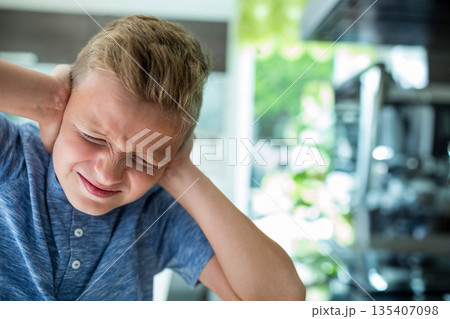 School age boy covering ears and squinting eyes in home kitchen with stainless steel range hood 135407098