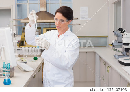 Female scientist at lab bench wearing lab coat pouring blue solution with pipette into test tube 135407108
