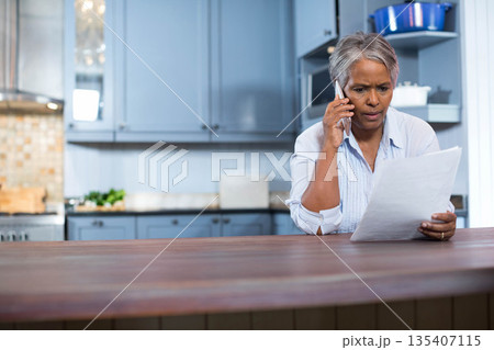 Senior African American woman holding smartphone, reading document at kitchen island, copy space 135407115