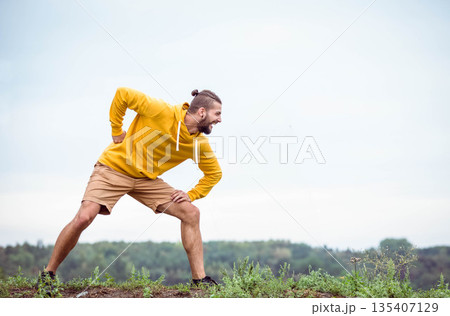 Male wearing yellow hoodie shorts running shoes doing side lunge stretch on grassy hilltop 135407129