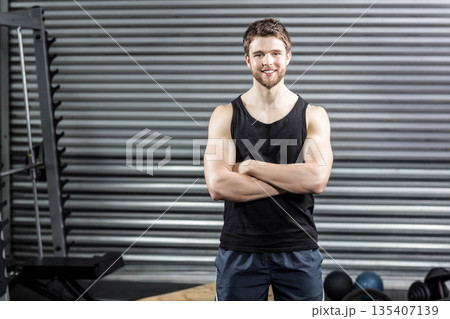 Muscular man folding arms and smiling toward camera inside gym area with squat rack and kettlebells Muscular man folding arms and smiling toward camera inside gym area with squat rack and kettlebells 135407139