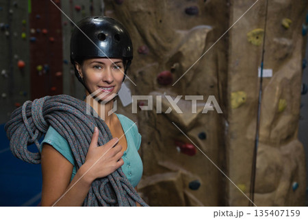 Female climber standing in climbing gym with rope near rock wall wearing helmet, copy space Female climber standing in climbing gym with rope near rock wall wearing helmet, copy space 135407159