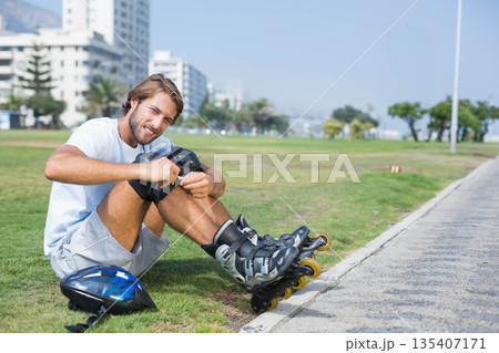 Male fastening knee pad beside park walkway, inline skates, wrist guards, helmet, copy space Male fastening knee pad beside park walkway, inline skates, wrist guards, helmet, copy space 135407171