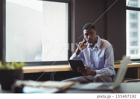 African American man sitting on bench by window in office holding tablet near laptop African American man sitting on bench by window in office holding tablet near laptop 135407192