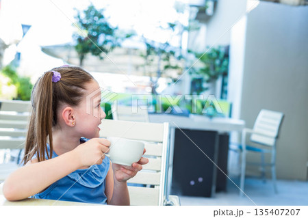 Female child holding white mug, sitting at table on cafe terrace among potted plants, copy space 135407207