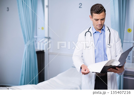 male doctor standing beside privacy curtains in cubicle reviewing file with stethoscope, copy space male doctor standing beside privacy curtains in cubicle reviewing file with stethoscope, copy space 135407216
