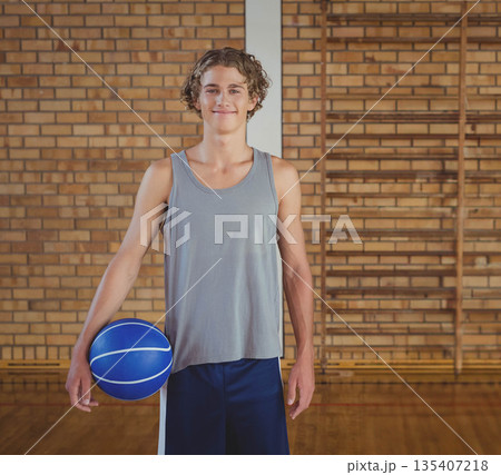 Teenage boy athlete smiling while holding blue basketball against brick wall in school gym 135407218