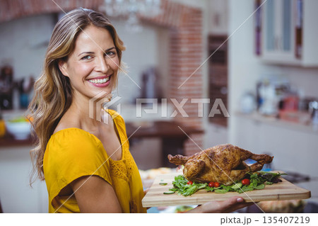 Woman in her thirties holding cutting board with roasted chicken in home kitchen with brick oven Woman in her thirties holding cutting board with roasted chicken in home kitchen with brick oven 135407219