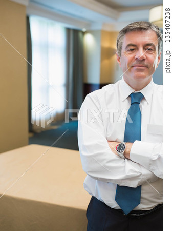 Middle-aged man standing with arms crossed in hotel conference room with tablecloth, copy space Middle-aged man standing with arms crossed in hotel conference room with tablecloth, copy space 135407238