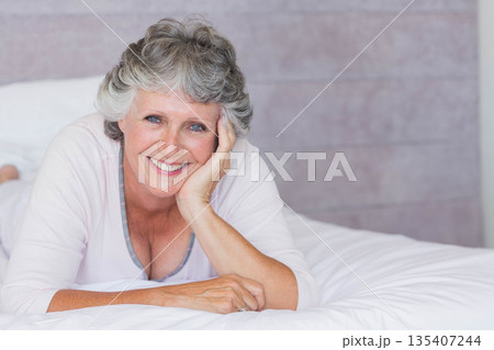 Senior woman lying on stomach on bed, wearing pink sleepwear and showing ring against wooden wall 135407244