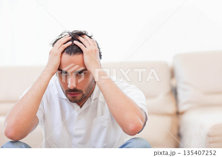 Man leaning forward in living room gripping head by sofa in polo shirt and jeans 135407252