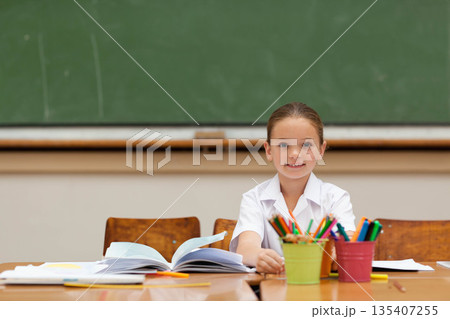 Female child student studying at wooden desk in classroom with open workbook, pencils and pens 135407255