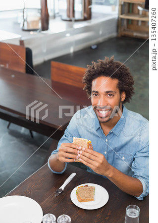 African American man smiling while holding whole-grain sandwich on cafeteria table with water glass 135407260