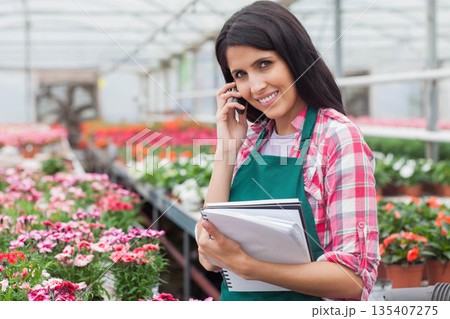 Woman greenhouse worker using notebook and talking on smartphone in potted plant aisles, copy space 135407275
