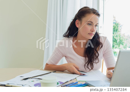 Female remote worker holding payment card while scanning laptop screen at home desk with coffee cup Female remote worker holding payment card while scanning laptop screen at home desk with coffee cup 135407284
