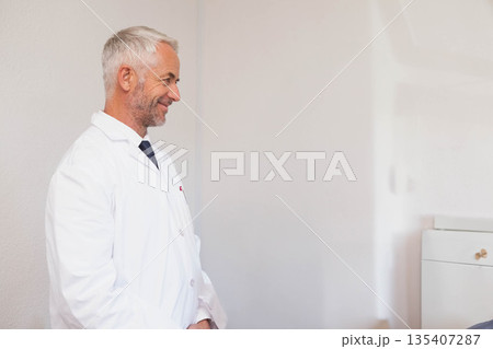 Senior male doctor wearing lab coat and tie standing in clinic beside white cabinet, copy space Senior male doctor wearing lab coat and tie standing in clinic beside white cabinet, copy space 135407287