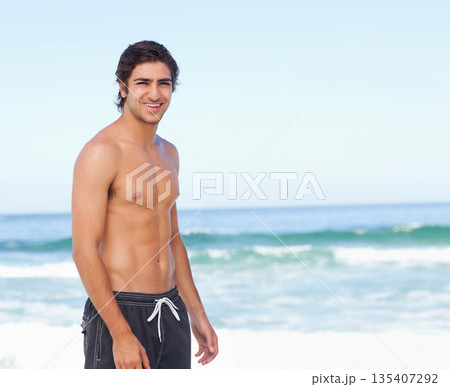 Shirtless man standing on sandy beach at water's edge wearing dark swim trunks amid ocean waves Shirtless man standing on sandy beach at water's edge wearing dark swim trunks amid ocean waves 135407292