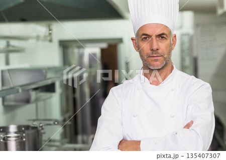 Mature male chef standing with crossed arms in professional kitchen with pots and pans, copy space 135407307