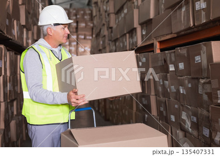 Male warehouse worker lifting cardboard box onto hand truck in warehouse aisle in reflective vest Male warehouse worker lifting cardboard box onto hand truck in warehouse aisle in reflective vest 135407331