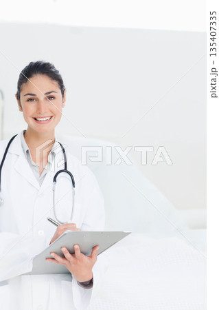 Female doctor standing beside bed with stethoscope holding clipboard and pen in clinic, copy space Female doctor standing beside bed with stethoscope holding clipboard and pen in clinic, copy space 135407335