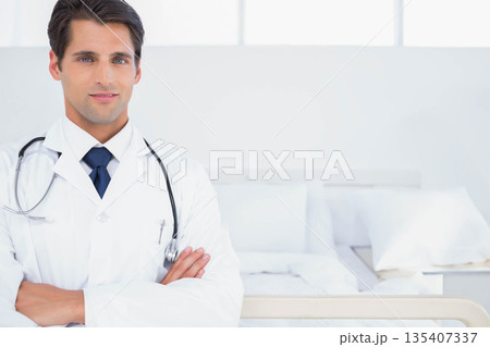 Male doctor in lab coat standing crossing arms beside hospital bed with stethoscope, copy space Male doctor in lab coat standing crossing arms beside hospital bed with stethoscope, copy space 135407337