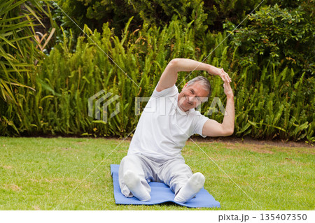 Senior male in sportswear sitting on blue exercise mat on grass lawn in garden stretching muscles 135407350