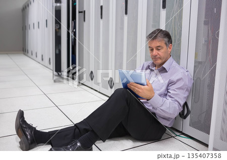 Senior man in dress clothes leaning against server rack, checking tablet at data center, copy space 135407358