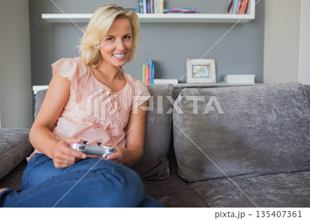 Adult woman sitting on gray sofa at home holding game controller smiling by bookshelf, copy space Adult woman sitting on gray sofa at home holding game controller smiling by bookshelf, copy space 135407361