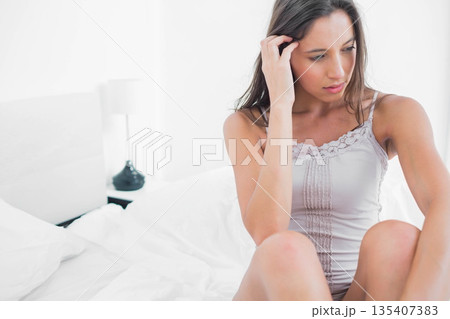 Woman sitting on white bedding in bedroom touching hair and gazing down near lamp, copy space Woman sitting on white bedding in bedroom touching hair and gazing down near lamp, copy space 135407383