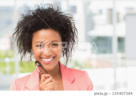 Smiling African American woman in coral blazer holding eyeglasses at office window over skyline Smiling African American woman in coral blazer holding eyeglasses at office window over skyline 135407384
