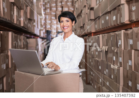 Asian woman working in warehouse aisle wearing button-down shirt at box workstation with laptop Asian woman working in warehouse aisle wearing button-down shirt at box workstation with laptop 135407392