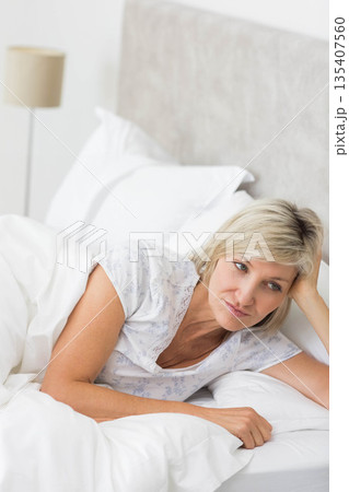 Mature woman lying on bed under gray upholstered headboard in bedroom wearing floral pajama top Mature woman lying on bed under gray upholstered headboard in bedroom wearing floral pajama top 135407560