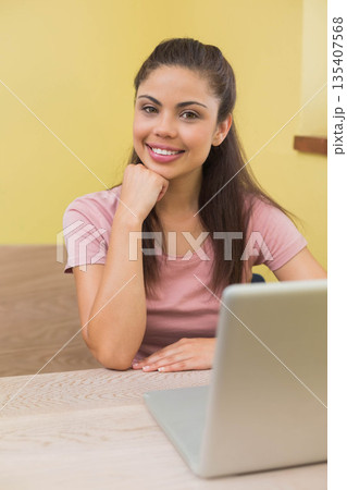 Woman sitting table at home using silver laptop, resting chin on fist and smiling toward viewer Woman sitting table at home using silver laptop, resting chin on fist and smiling toward viewer 135407568
