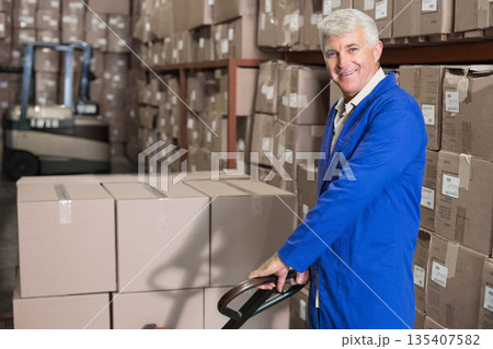 Middle-aged man in coat operating pallet truck carrying boxes in warehouse rack aisle, copy space 135407582