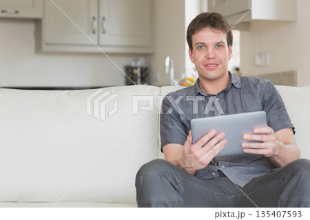 Mid adult man wearing grey shirt sitting on couch holding tablet with kitchen cabinets, copy space 135407593