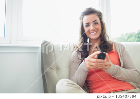 Woman holding smartphone while sitting on beige sofa by large window in living room, copy space Woman holding smartphone while sitting on beige sofa by large window in living room, copy space 135407594