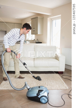 Man vacuuming area rug with vacuum cleaner on tiled floor in living room near kitchen cabinets Man vacuuming area rug with vacuum cleaner on tiled floor in living room near kitchen cabinets 135407595