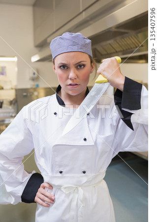 Female chef wearing checkered hat holding large kitchen knife near face in commercial kitchen 135407596
