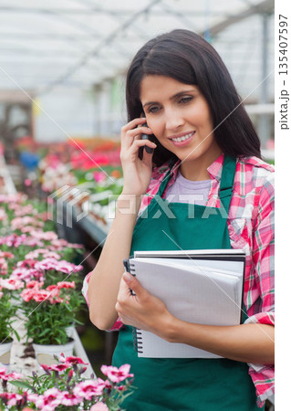 Female horticultural worker talking on smartphone while holding notebooks in greenhouse, copy space 135407597